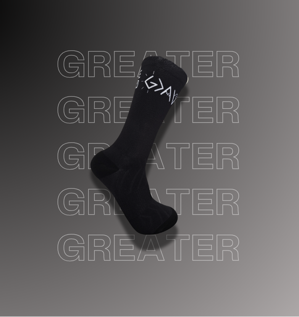 A single black crew sock with a white God is greater than my highs and lows symbol, shown floating against a dark gray gradient background with repeated faint 'GREATER' text and website watermarks.