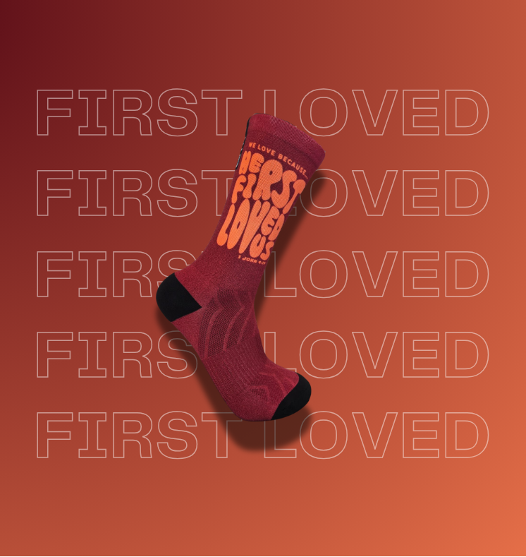 A single crew sock floating on a warm orange gradient background with repeated faint 'FIRST LOVED' text and website watermarks. The sock is dark red with black toe and heel areas and large orange text reading 'HE FIRST LOVED US' on the side.
