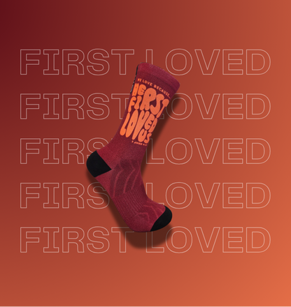 A single crew sock floating on a warm orange gradient background with repeated faint 'FIRST LOVED' text and website watermarks. The sock is dark red with black toe and heel areas and large orange text reading 'HE FIRST LOVED US' on the side.