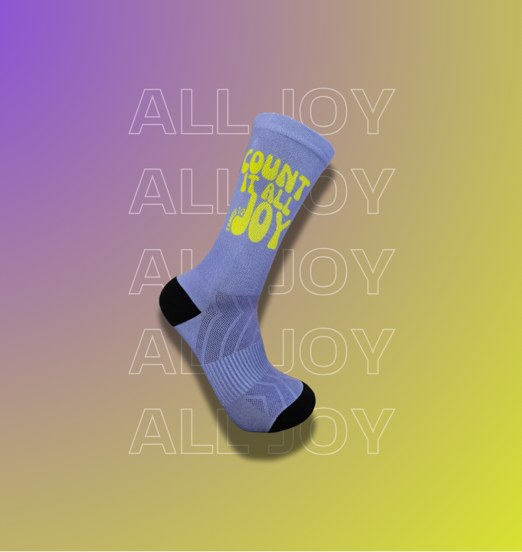 A single crew sock floating on a purple-to-yellow gradient background. The sock is light blue with a bright yellow 'COUNT it all JOY' text on the side and black toe/heel areas.