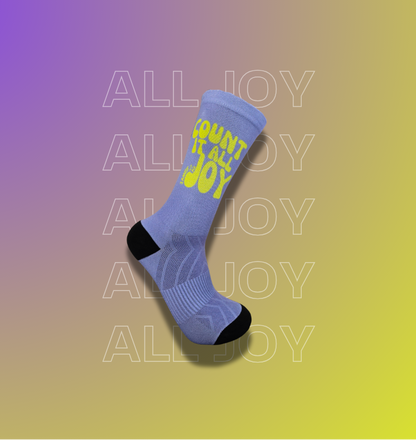 A single crew sock floating on a purple-to-yellow gradient background. The sock is light blue with a bright yellow 'COUNT it all JOY' text on the side and black toe/heel areas.
